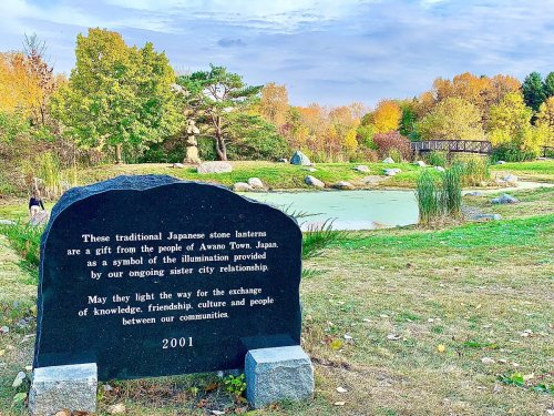 A stone lantern in a park, with a plaque about its significance, surrounded by autumn foliage and a pond.
