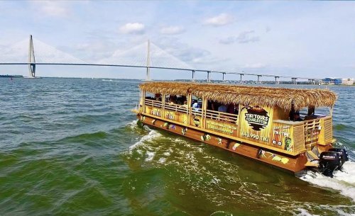 A colorful tour boat with a thatched roof navigates a river under a bridge on a sunny day.
