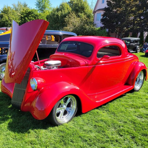 A shiny red classic car with an open hood, parked on green grass at a car show.