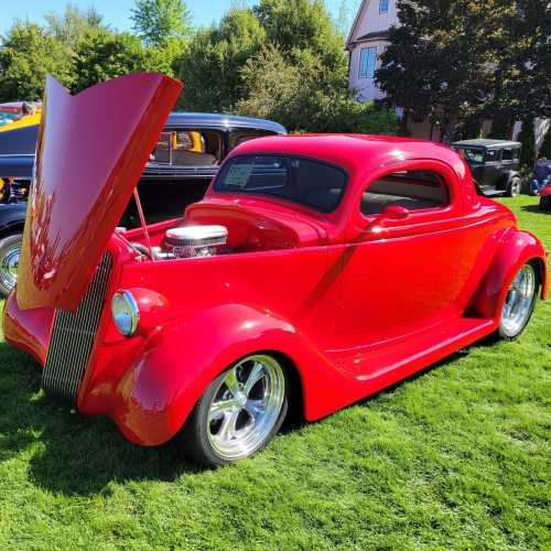 A shiny red classic car with an open hood, parked on green grass at a car show.