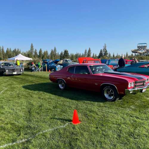 A classic red car parked on grass at a car show, with other vintage vehicles and trees in the background.