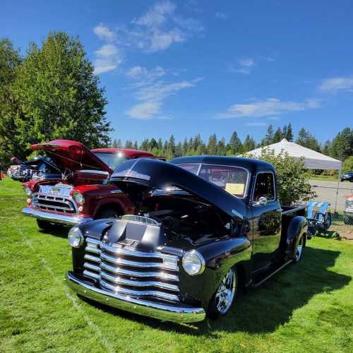 A classic black pickup truck with the hood open, parked on grass, with another vintage truck in the background under a blue sky.