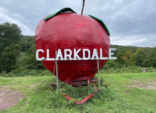 A large, red strawberry sculpture with the word "CLARKDALE" in white letters, set against a cloudy sky and green landscape.