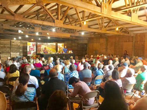 A large audience sits in a rustic barn, listening to speakers at a floral event, surrounded by colorful displays.