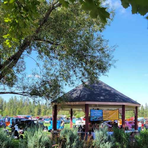 A gazebo surrounded by classic cars at an outdoor event, with trees and a clear blue sky in the background.