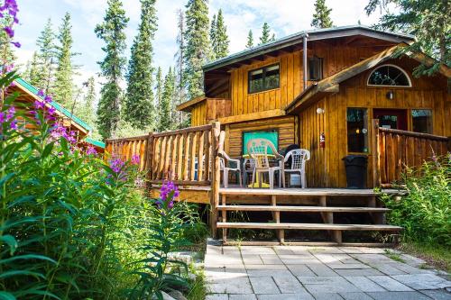 A wooden cabin surrounded by trees and flowers, featuring a deck with chairs and a welcoming entrance.