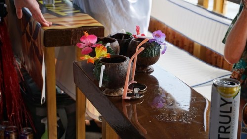 Three tropical drinks with colorful flowers and straws sit on a wooden bar, with a can of beverage nearby.