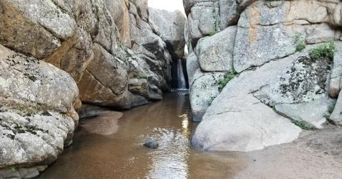 A narrow canyon with towering rock formations and a small stream flowing through the sandy bottom.