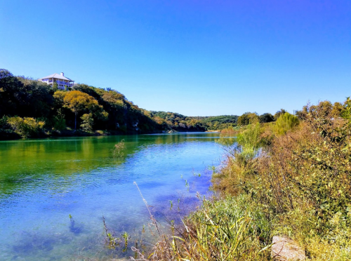 A serene river scene with lush greenery and a house on a hill under a clear blue sky.