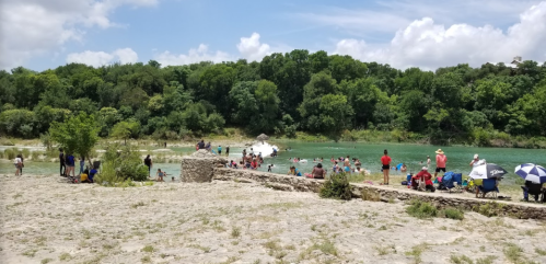 A sandy area by a river with people swimming and relaxing under trees, enjoying a sunny day.