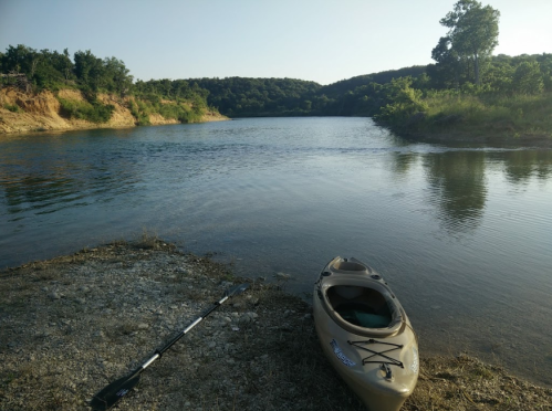 A calm river scene with a kayak and paddle on the shore, surrounded by lush greenery and hills in the background.