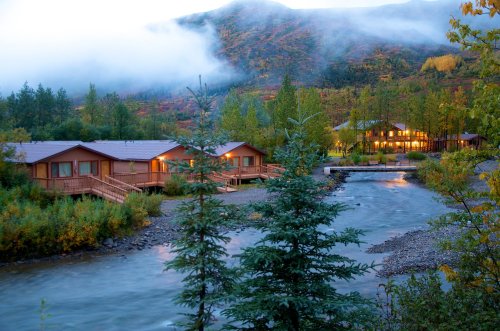 A serene riverside scene featuring wooden cabins surrounded by trees and misty mountains at dawn.