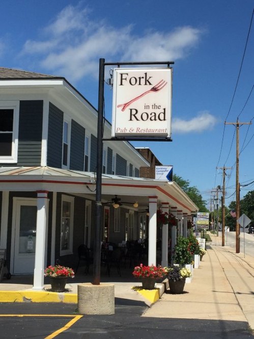 Sign for "Fork in the Road Pub & Restaurant" outside a building, with flowers and a clear blue sky in the background.