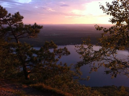 A serene landscape at sunset, featuring a river surrounded by trees and distant mountains under a colorful sky.