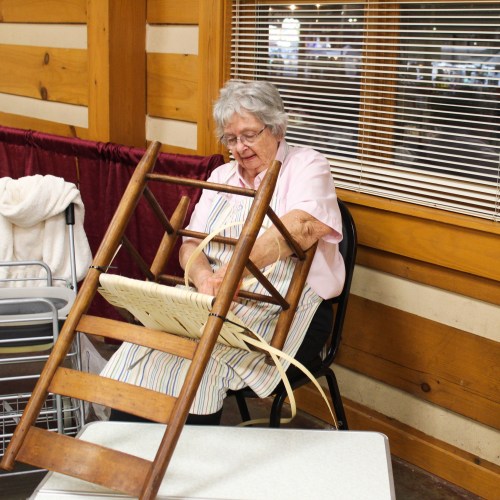An elderly woman repairs a wooden chair in a cozy, rustic setting with wooden walls and large windows.