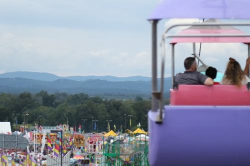A couple enjoys a scenic view from a ferris wheel overlooking a vibrant fairground with rides and colorful tents.