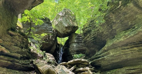 A rocky landscape with a large boulder balanced between two cliffs, surrounded by lush green foliage and a small waterfall.