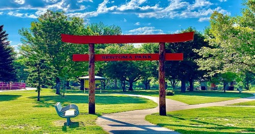 A red torii gate marks the entrance to Sertoma Park, surrounded by lush greenery and a clear blue sky.