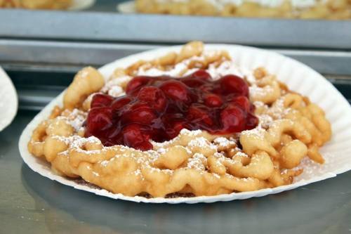 A golden funnel cake topped with cherry filling and powdered sugar on a white paper plate.