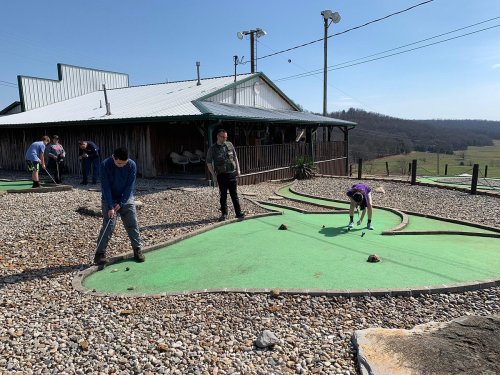 A mini-golf course with players putting on green turf, surrounded by gravel and a rustic building in the background.