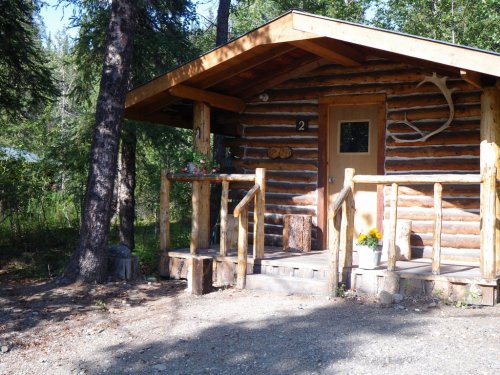A rustic log cabin with a wooden porch, surrounded by trees and greenery.