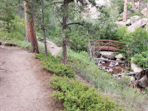 A winding dirt path leads to a wooden bridge over a small stream, surrounded by trees and rocky terrain.