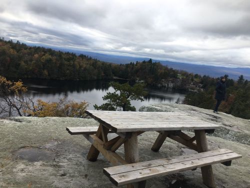 A wooden picnic table overlooks a serene lake surrounded by trees under a cloudy sky. A person stands in the distance.