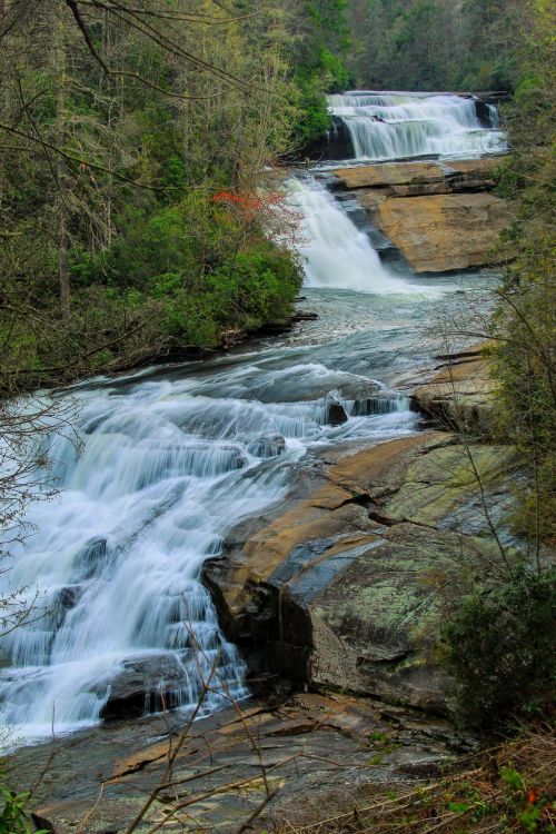 A serene waterfall cascades over rocky ledges, surrounded by lush greenery and trees in a tranquil natural setting.