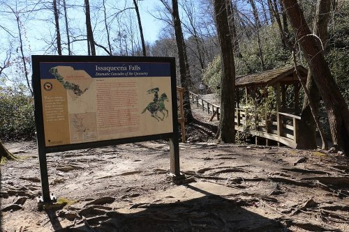 Sign for Issaqueena Falls in a wooded area, with a nearby wooden bridge and trail visible in the background.