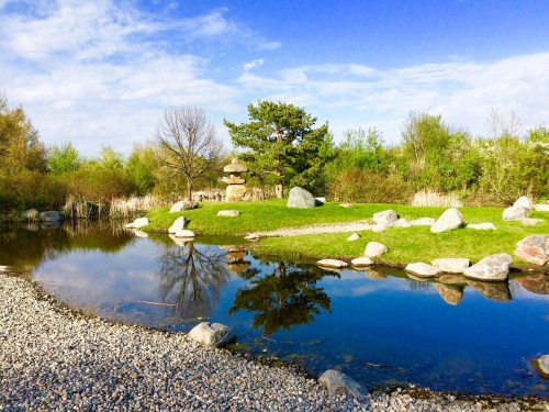 A serene landscape featuring a pond, green grass, rocks, and trees under a blue sky with scattered clouds.
