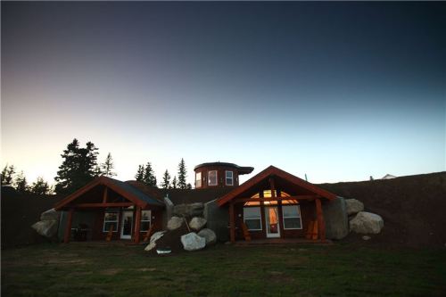 A unique cabin with wooden architecture, surrounded by large rocks and trees, under a twilight sky.