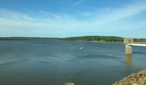 A calm lake surrounded by green hills under a blue sky, with a small boat near a concrete dock.