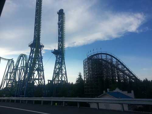 Two tall roller coasters and a wooden coaster against a blue sky with clouds, surrounded by trees.