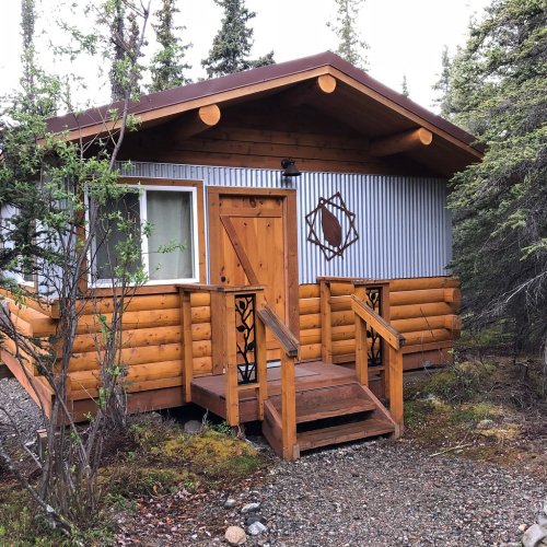 A rustic cabin with wooden logs and a metal exterior, surrounded by trees and gravel.