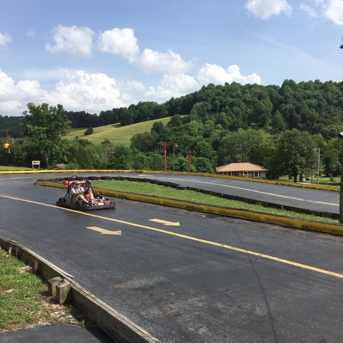 A go-kart track with two karts racing on a sunny day, surrounded by green hills and trees.
