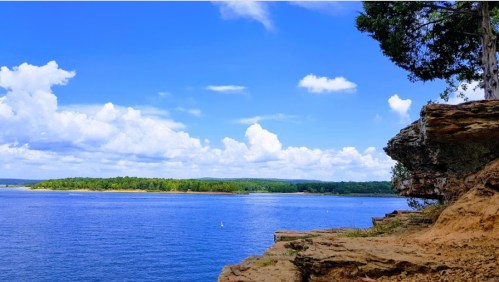 A scenic view of a calm blue lake surrounded by green trees and a rocky shoreline under a bright blue sky.