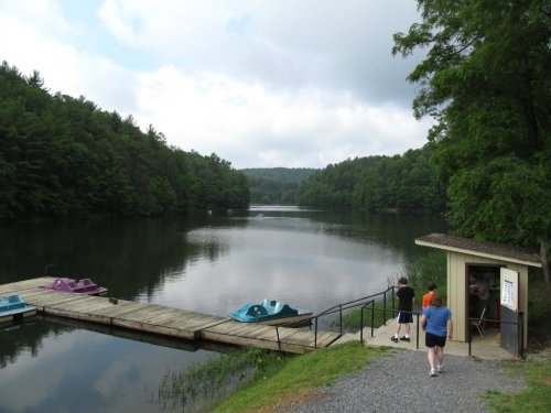 A serene lake surrounded by trees, with a dock and colorful paddle boats, under a cloudy sky. People walk nearby.
