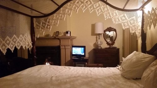 A cozy bedroom with a canopy bed, a fireplace, a TV, and a vintage dresser reflecting soft lighting.