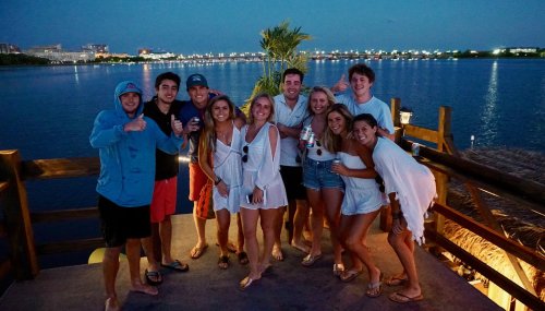 A group of ten friends poses together on a deck by the water at dusk, smiling and enjoying their time.