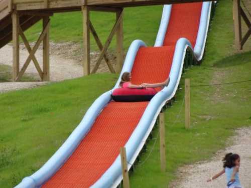 A child slides down a large, colorful water slide while another child runs nearby on a grassy area.