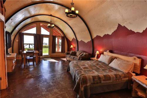 Cozy interior of a rustic room with a bed, seating area, and large windows showcasing a scenic view.