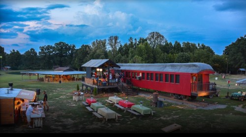 A red train car converted into a dining space, surrounded by picnic tables and a rustic outdoor setting at dusk.