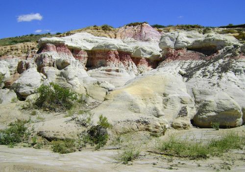 Colorful rock formations with white, pink, and yellow hues under a blue sky, surrounded by sparse vegetation.