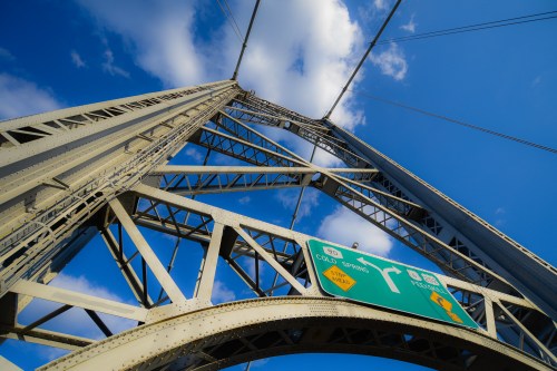 A low-angle view of a metal bridge structure against a blue sky with clouds, featuring a directional road sign.