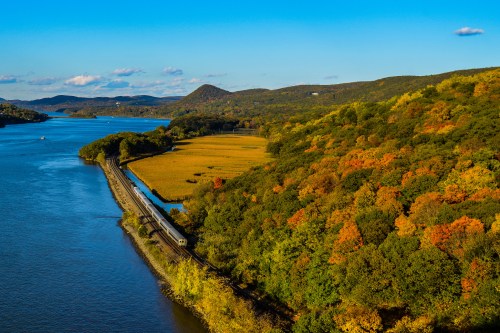 A scenic view of a river bordered by colorful autumn foliage and a train track along the water's edge.