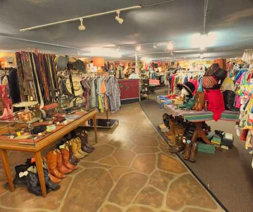 A vintage thrift store interior with clothing racks, boots, and accessories displayed on tables and shelves.