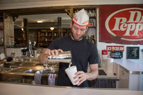 A man in a paper hat pours a milkshake from a metal shaker into a cup at a retro-style diner.