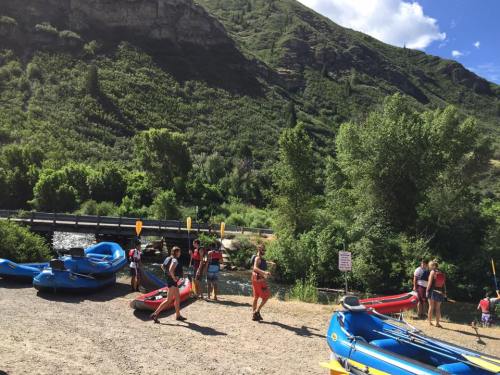 People preparing kayaks by a river, surrounded by lush greenery and a bridge in the background on a sunny day.