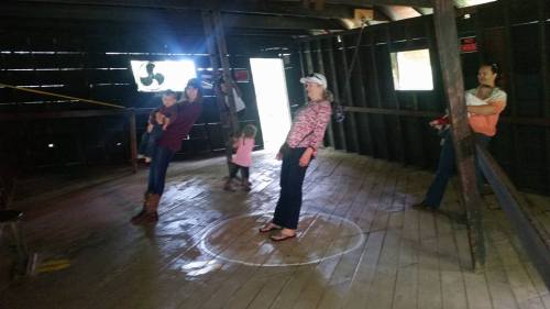 A group of people inside a rustic barn, some playing and others observing, with wooden beams and a circular marking on the floor.