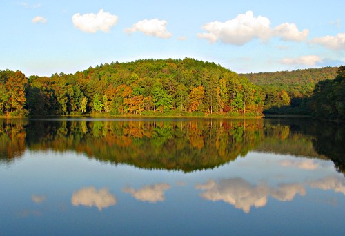 A serene lake reflecting a colorful forest and blue sky with fluffy clouds, creating a peaceful natural scene.